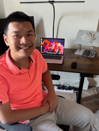 Young man, smiling while sitting at a desk with a laptop, screen and associated cables
