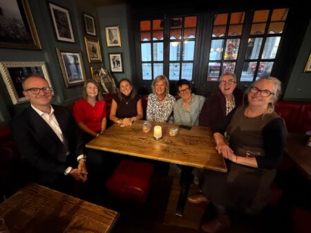 A group of people, sat around a dark wooden table all smiling at the camera.